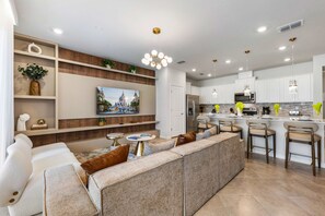 Un salon moderne avec un canapé d’angle, un téléviseur fixé au mur et un coin cuisine doté d’un îlot central et de tabourets de bar.