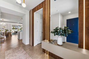 A modern interior with a hallway leading to a dining area, a reception desk with a plant, and a blue door. - Brand New | Modern 5BR with Pool & Game Room 16212 (Clermont)