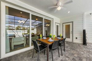 A covered patio with a table set for dining, a ceiling fan, and a sliding glass door leading to an outdoor area. - Brand New | Modern 5BR with Pool & Game Room 16212 (Clermont)