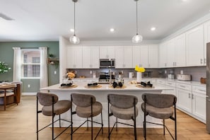 A modern kitchen with white cabinets, a central island with bar stools, and stainless steel appliances. - Brand New | Family 6BR Pool Spa & Game Lounge 7875 (Kissimmee)