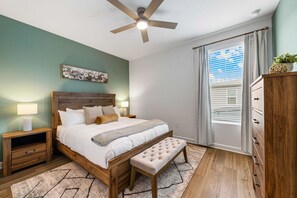 Ein Schlafzimmer mit einem Bett, Nachttischen, einer Bank, einem Deckenventilator, einem Fenster mit Jalousien und einer Kommode.