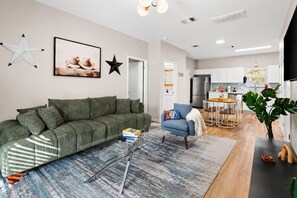A modern living room with a green sofa, a glass coffee table, and a blue armchair. There is a kitchen in the background with white cabinets and a dining area. - East Austin 4-Bedroom Home, Private Backyard with Plunge Pool & BBQ, Sleeps 11 (Austin)