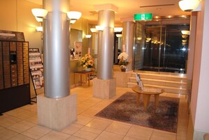 Um saguão de hotel com balcão de recepção, sinalização de saída verde e escada que leva a um andar superior.