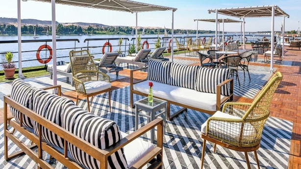 A terrace with outdoor seating, including sofas and chairs, overlooking a body of water with boats in the distance. - Movenpick MS Hamees (Qena)