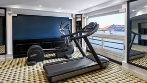 A modern gym with treadmills, a stationary bike, and a rowing machine, featuring a large window with a view of a body of water and hills. - Movenpick MS Hamees (Qena)