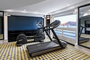 Ein modernes Fitnessstudio mit Laufbändern, einem Ergometer und einem Rudergerät, mit einem großen Fenster und Blick auf ein Gewässer und Hügel.