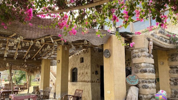 An outdoor seating area with tables and chairs, a stone wall with a carved tree trunk, and a covered pergola with hanging lights. - Zad El Mosafer (Yusuf as-Sidiq)