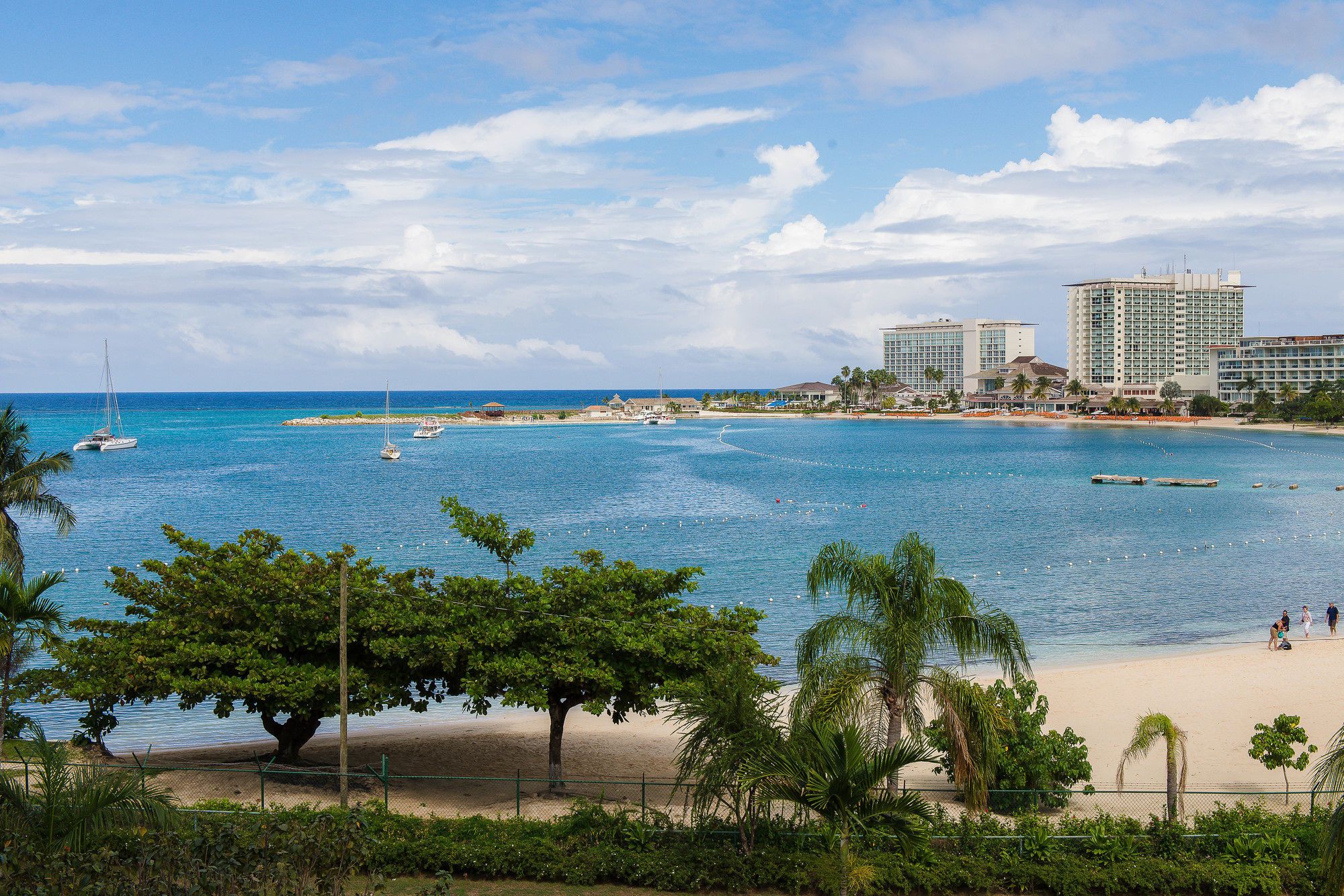 Vista costeira com praia arenosa, mar azul cristalino e alguns barcos.