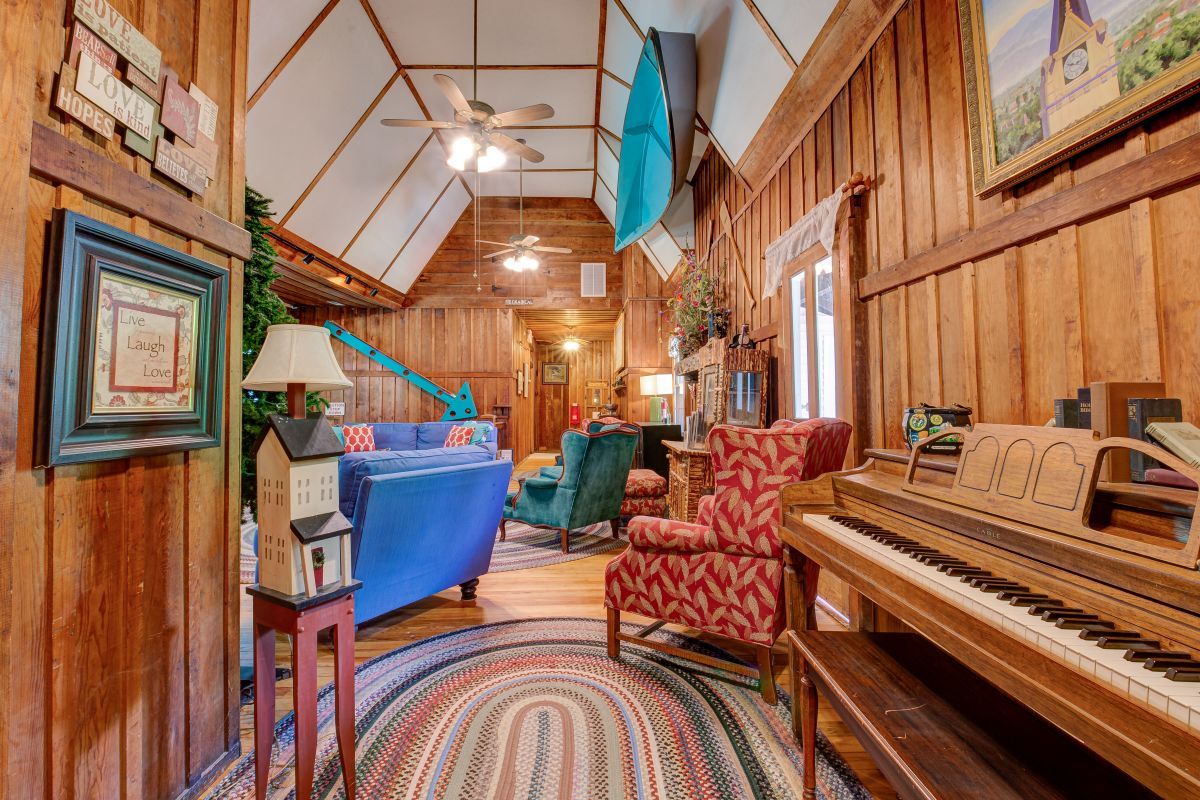 A cozy room with a wooden interior, a blue sofa, a red patterned armchair, a wooden piano, and a ceiling fan.