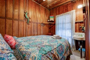 A wooden-paneled room with a bed, a hanging lizard, a painting, a window with curtains, and a sink.