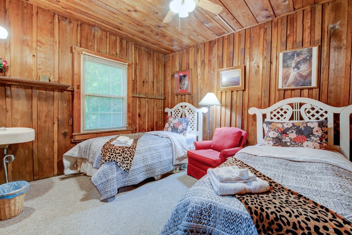A wooden-paneled room with two beds, a red chair, a lamp, and a window with blinds.