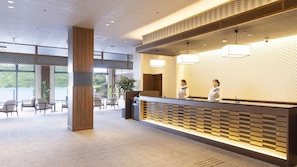 Zona de recepción con mostrador de madera, dos recepcionistas y vista al exterior a través de amplios ventanales.