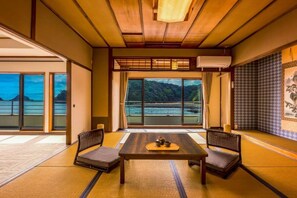 Una habitación japonesa tradicional con suelo de tatami, una mesa baja con cojines y vistas a un cuerpo de agua y colinas.