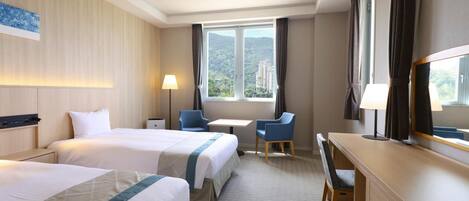 A hotel room with two beds, a desk, a chair, a lamp, and a large window with a view.