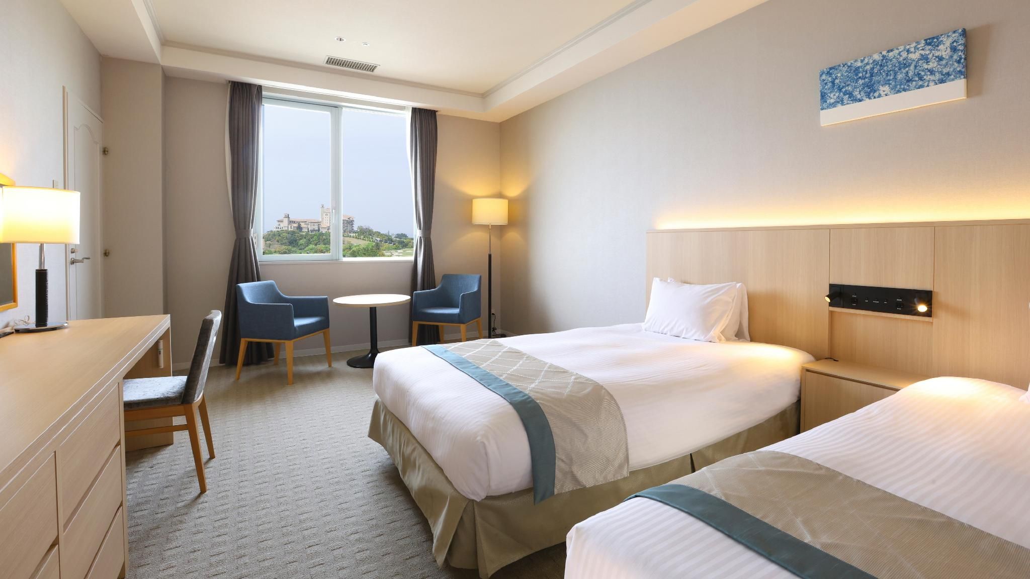 Habitación de hotel con dos camas, un escritorio, una silla, una lámpara y vista a un edificio a través de la ventana.