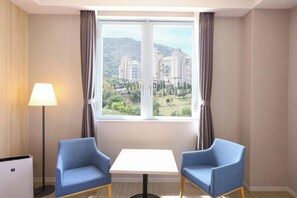 Une chambre d’hôtel avec une grande fenêtre, deux fauteuils bleus, une petite table blanche et un lampadaire.