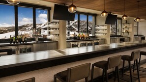 A bar area with a long counter, multiple seating options, and a view of a snowy mountain landscape through large windows.