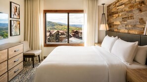 Une chambre d’hôtel avec un grand lit, une commode en bois, un banc, une fenêtre donnant sur des bâtiments et des collines, et un mur en bois.