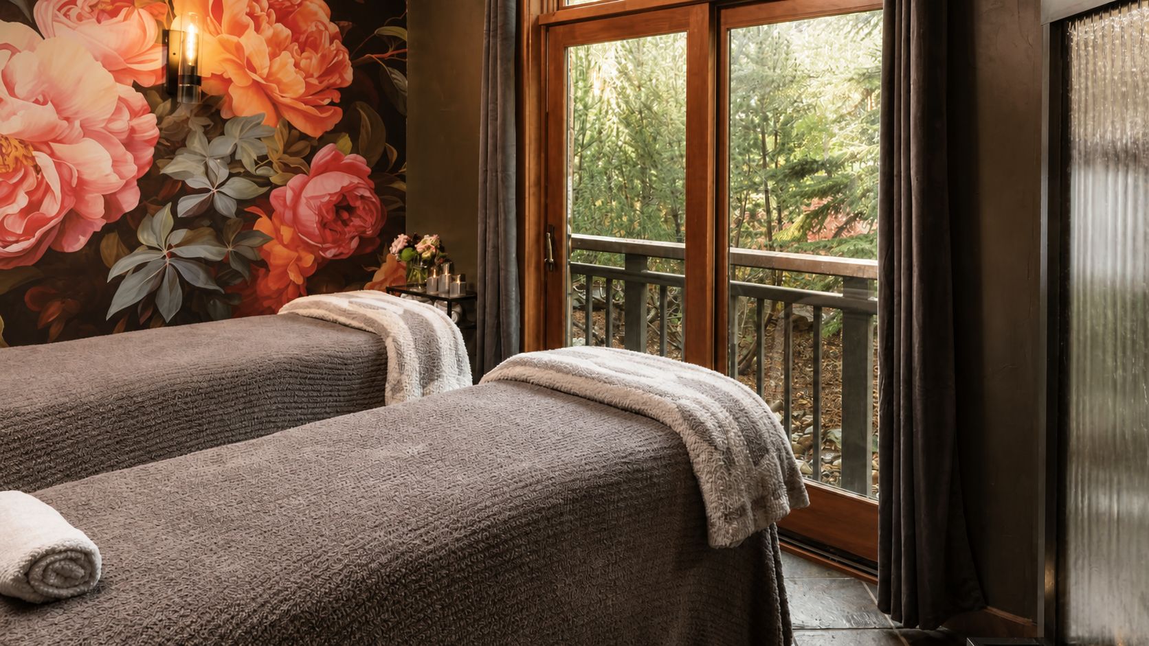 Ein Raum mit zwei Massagetischen, einer großen Blütenwand und einem Balkon mit Blick auf Bäume.