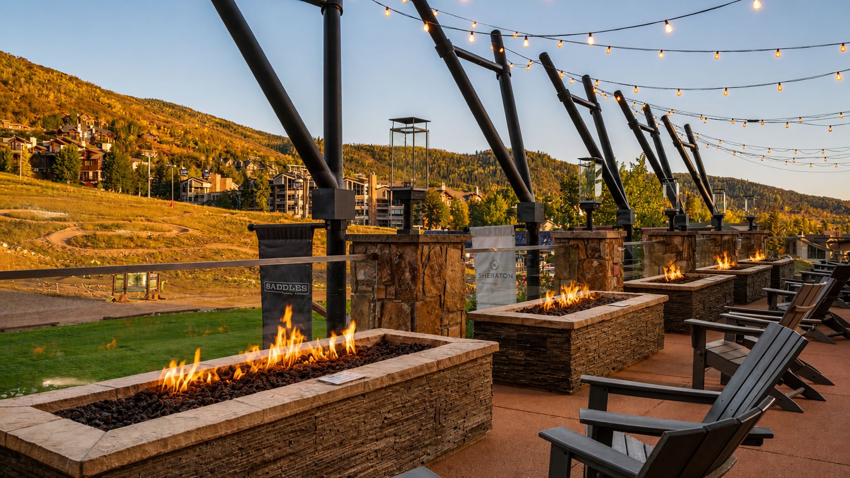 Outdoor seating with fire pits and string lights, overlooking a hilly landscape.