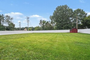 A grassy field with a white fence, a red building, and trees in the background.