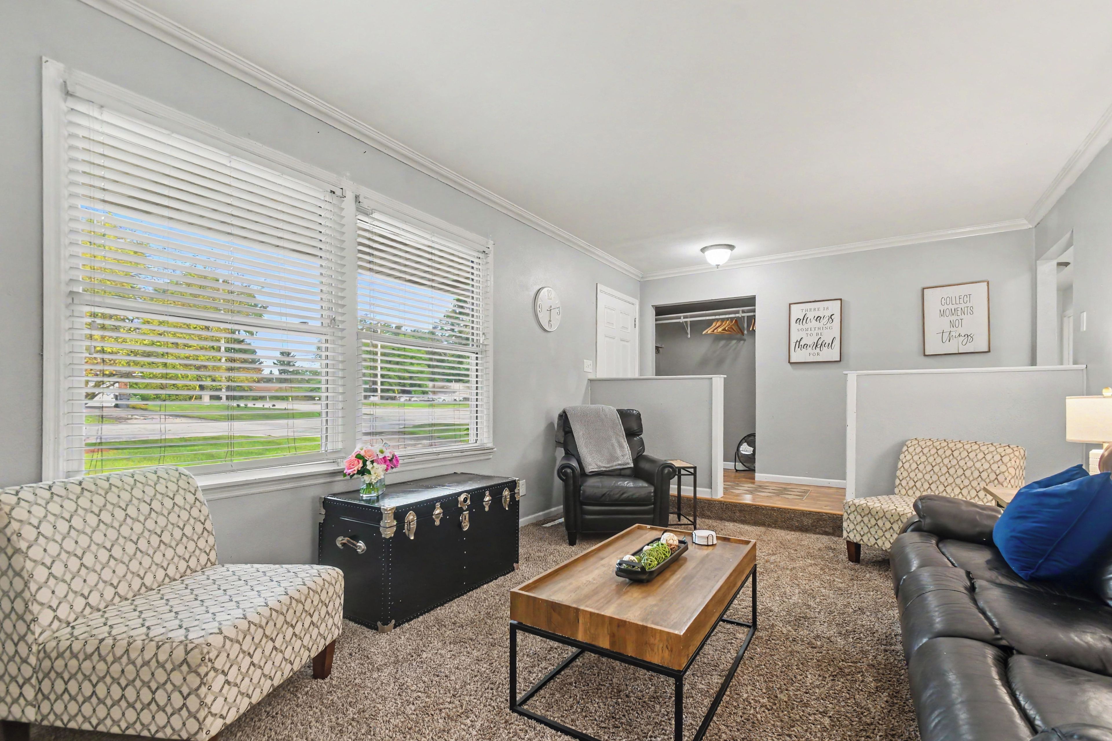 A living room with a sofa, a coffee table, a black suitcase, and a chair. There are two windows with blinds, a door leading to a closet, and a wall with framed inspirational quotes.