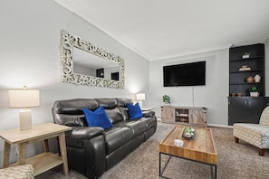 A living room with a black leather sofa, two blue cushions, a wooden coffee table, a TV mounted on the wall, and a black shelving unit with decorative items.
