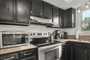 A modern kitchen with dark cabinetry, a stainless steel microwave, stove, and oven, a coffee maker, and a window with a view of trees. - Notre Dame Game Day Getaway! Minutes from Campus, South Bend & Eddy Street (South Bend)