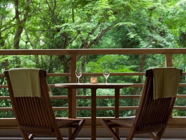 Una mesa de madera con dos sillas, una lámpara de mesa pequeña y vistas a un bosque a través de la barandilla de un balcón.