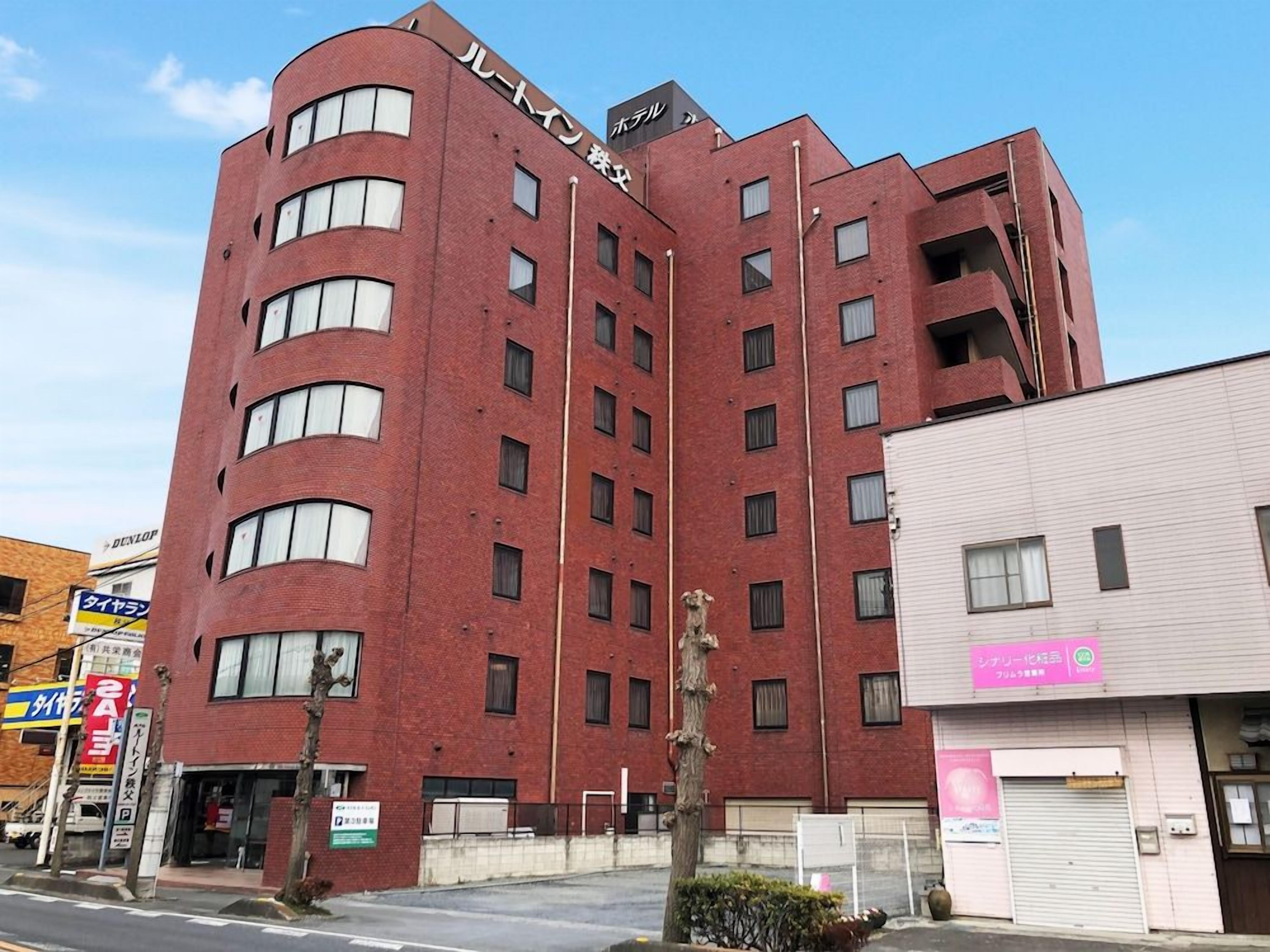 Un edificio de ladrillo rojo de varios pisos con una esquina curva, que presenta un letrero con caracteres japoneses, un escaparate visible con un letrero rosa y un árbol en frente.