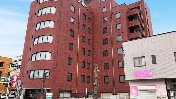 Edifício de tijolos vermelhos com vários andares e canto curvo, exibindo uma placa com caracteres japoneses, uma vitrine visível com uma placa rosa e uma árvore na frente.