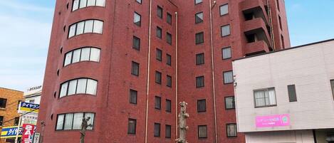 Un edificio de ladrillo rojo de varios pisos con una esquina curva, que presenta un letrero con caracteres japoneses, un escaparate visible con un letrero rosa y un árbol en frente.