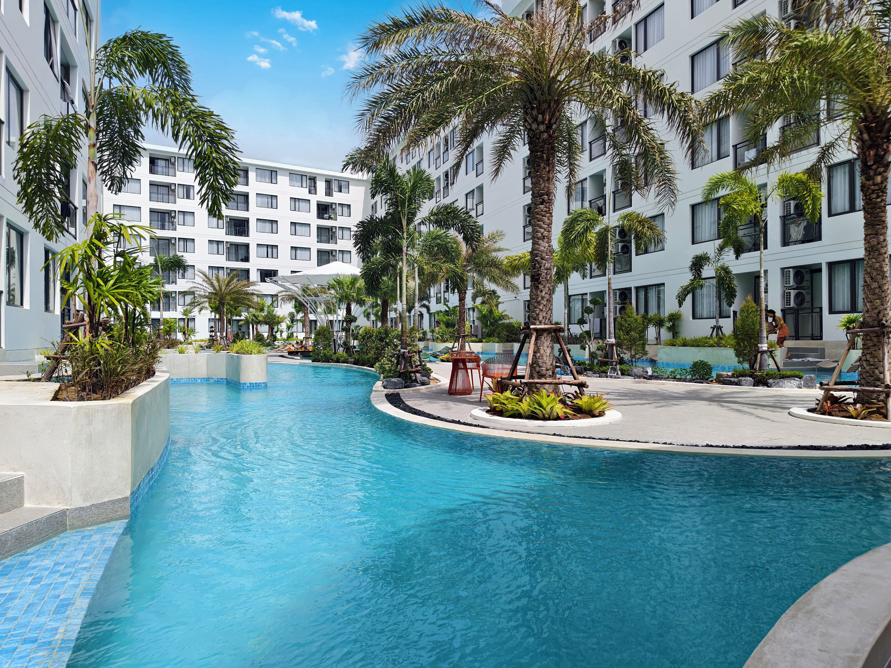 Un espace piscine avec une eau d’un bleu limpide, entouré de palmiers et bordé d’un immeuble à plusieurs étages doté de balcons.