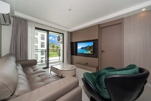 Habitació d'hotel moderna amb un sofà, una televisió penjada a la paret, un balcó amb vistes a edificis i vegetació, i un aire acondicionador fixat al sostre.