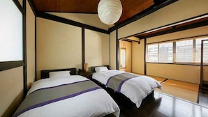 Quarto tradicional japonês com tatames, duas camas com roupa de cama branca e detalhes em roxo, um pendente de papel e uma porta deslizante que dá para outro cômodo.