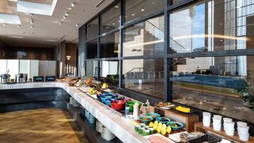 Zona de bufé con una variedad de alimentos y bebidas, una barra larga y vistas a la cocina a través de amplios ventanales.