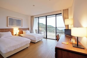 A hotel room with two beds, a desk, a TV, and a balcony with a view of the hills.