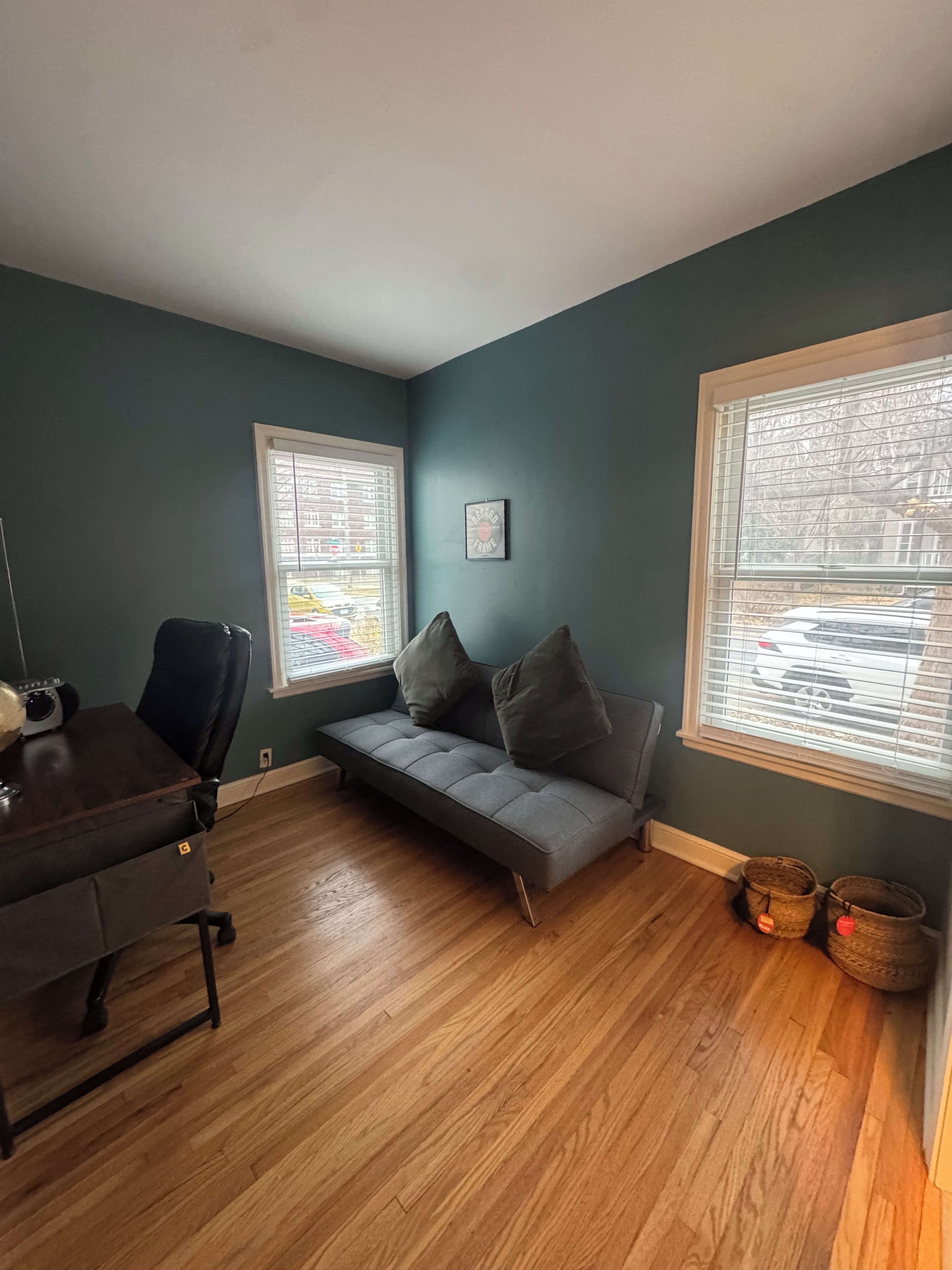 Une pièce avec un mur de couleur bleu-vert foncé, un canapé gris, un bureau en bois et une fenêtre avec des stores.