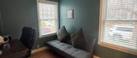 Una habitación con pared verde azul oscuro, un sofá gris, un escritorio de madera y una ventana con persianas.