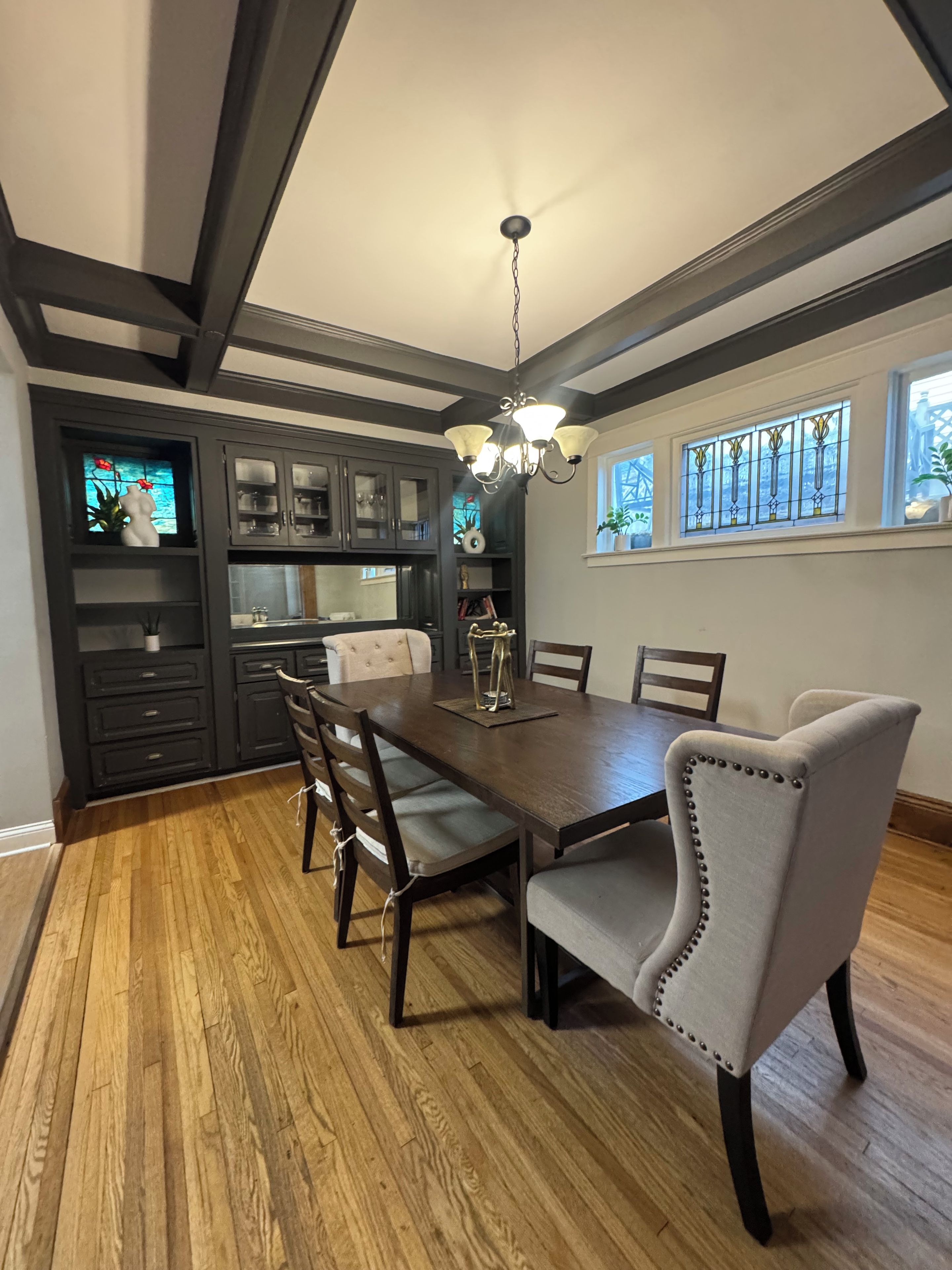 A dining area with a wooden table, six chairs, a chandelier, dark cabinetry, and a window with stained glass.