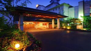 A hotel entrance with a covered walkway, a parking area, and a well-lit garden.