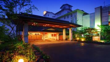 A hotel entrance with a covered walkway, a parking area, and a well-lit garden.
