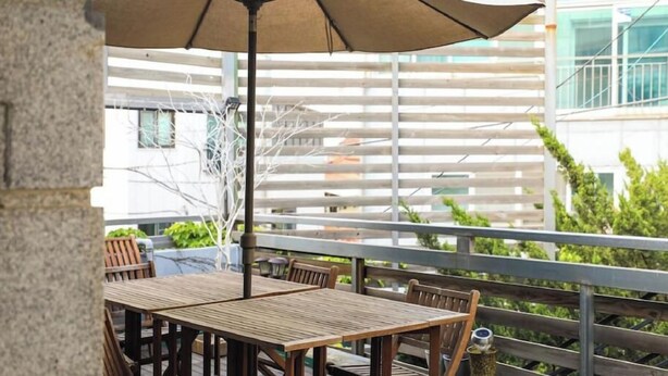 An outdoor patio with wooden tables and chairs, a metal railing, and a building in the background.