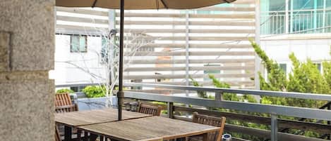 Une terrasse extérieure avec des tables et des chaises en bois, une rambarde en métal et un bâtiment en arrière-plan.
