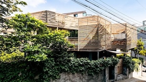 A modern residential building with a sloped roof, wooden paneling, and a stone wall with greenery.