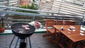 A rooftop patio with a grill, wooden table, chairs, and a potted plant.
