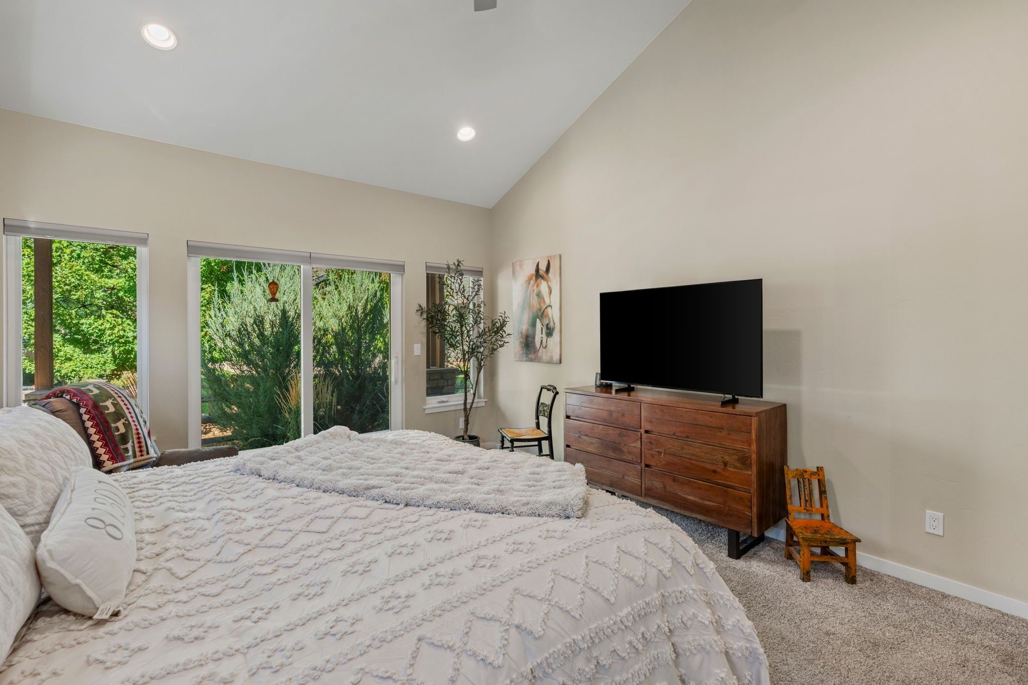 A bedroom with a bed, a dresser, a television, and a window with a view of greenery.