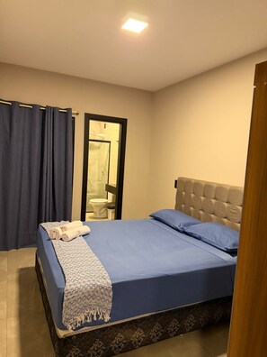 Une chambre à coucher avec un lit, une salle de bain visible à travers une porte et un luminaire au plafond.