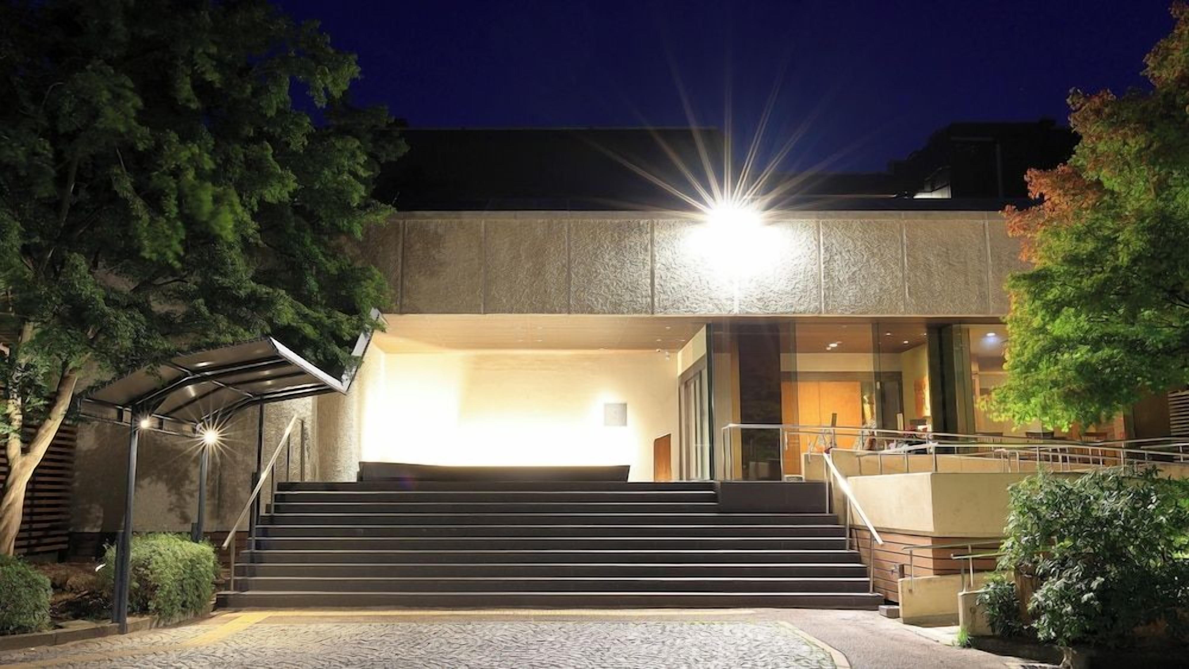 A building entrance with a staircase leading up to a covered area, surrounded by trees and a paved path.