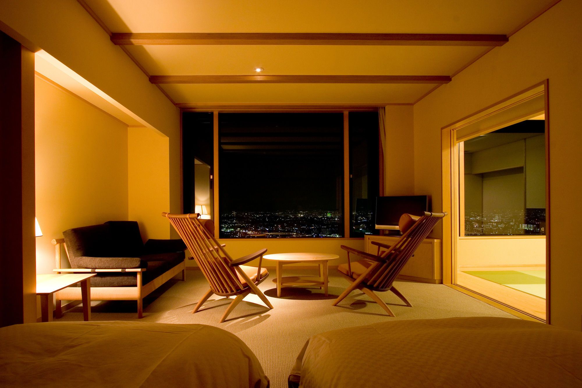 A hotel room with a bed, two chairs, a small table, and a large window offering a city view.